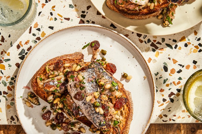 Two white plates of sardines on toast, scattered with raisins and pine nuts on a wooden table