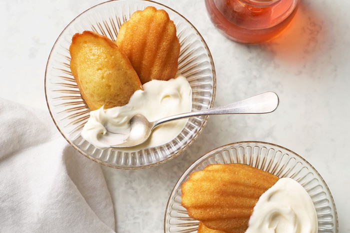 Two glass bowls with two madeleines and a dollop of cream next to a jug of syrup