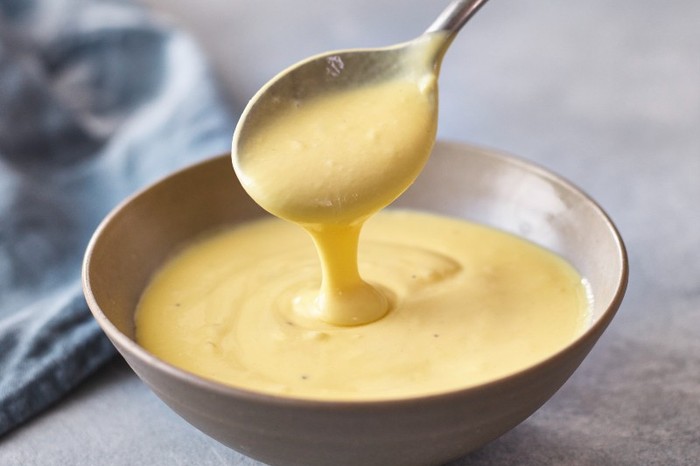 A small bowl of hollandaise with more sauce running off a silver spoon