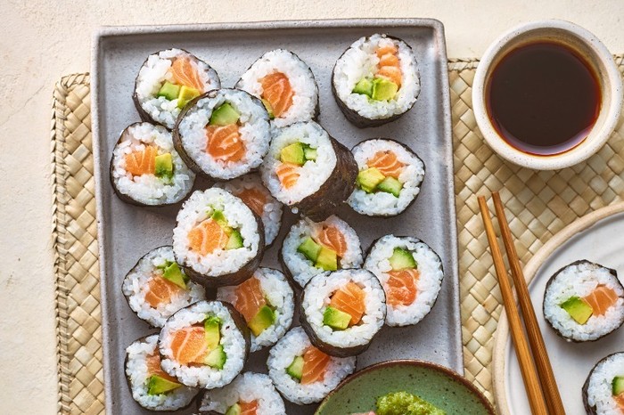 Sushi rolls on a grey rectangular platter with picked ginger and wasabi and a pot of soy sauce