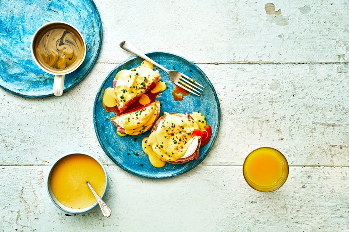 A white-washed background topped with blue plates, with two English muffins, poached eggs and hollandaise sauce on top. There is a cup of coffee and glass of orange juice on the table