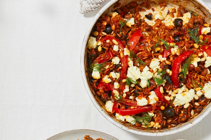 A casserole dish filled with tomato rice, red peppers and topped with pine nuts and feta