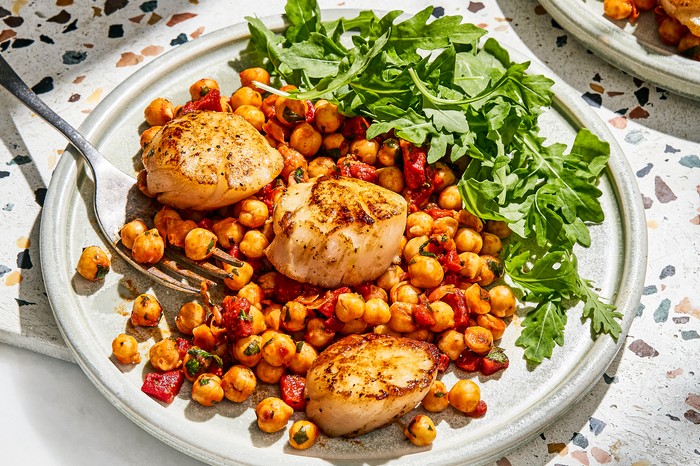 A white plate piled with three large scalops on a bed of golden chickpeas with a garnish of rocket to the side