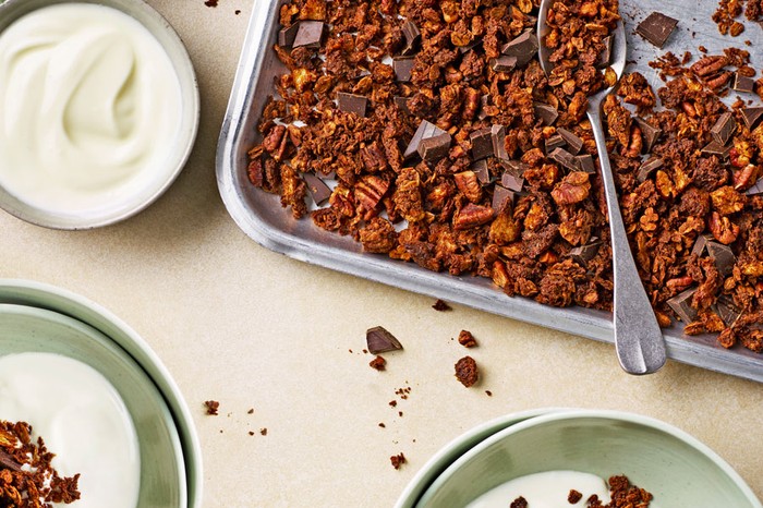 Baking tray of sourdough chocolate granola next to a bowl of yoghurt and two bowls of granola and yoghurt