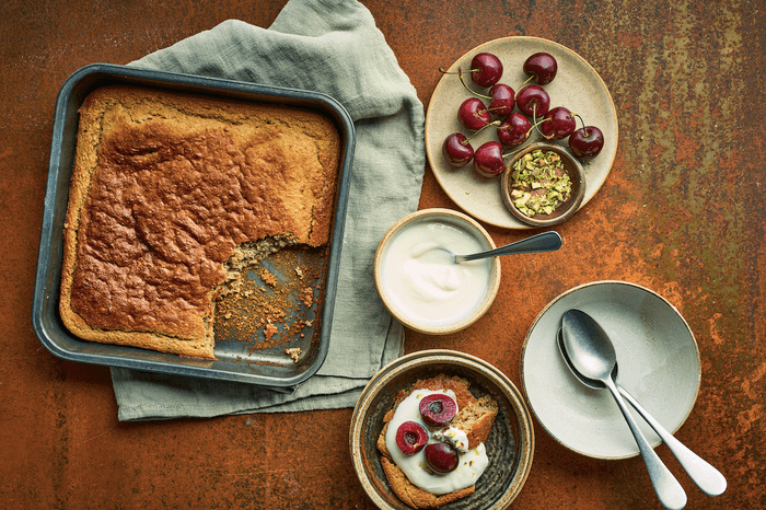 Baking tin filled with golden baked oats on a linen napkin next to a plate of cherries and chopped nuts and a bowl of yogurt