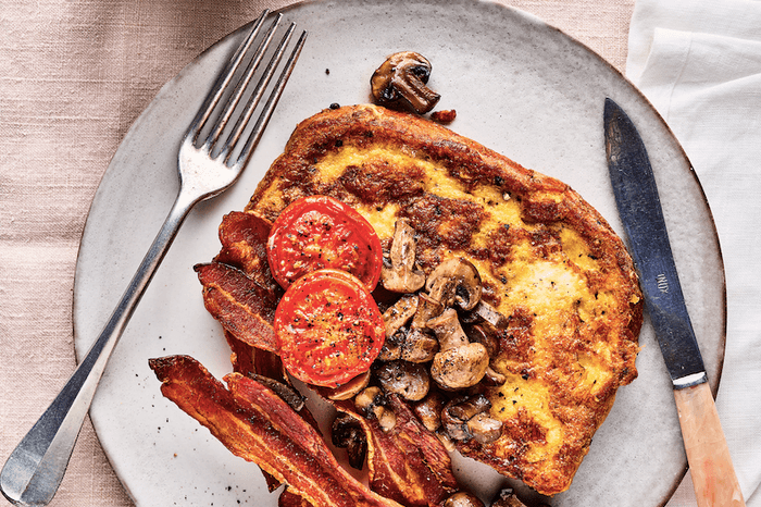 Eggy bread, tomatoes, mushrooms and bacon on a white plate with a knife, fork and cup of coffee, on a linen cloth