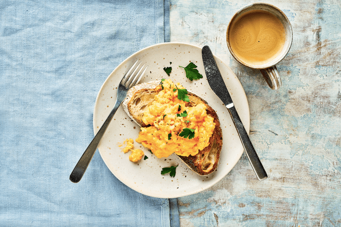 A plate of scrambled eggs on a slice of sourdough with a knife and fork and cup of coffee on a piece of blue fabric